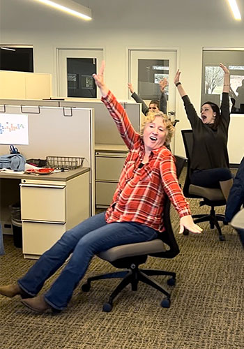 Three Love & Company employees sitting in their office chairs with their hands up on the air as part of a music video